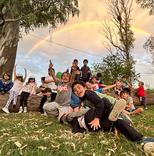 alumnos motrinfan de campamento, sentados en el pasto con el arcoiris de fondo